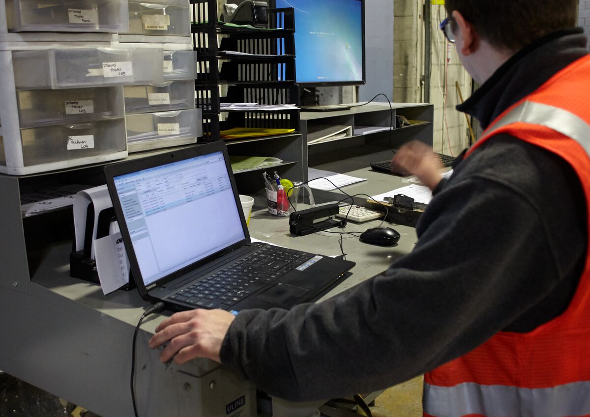 Person in a safety vest working at a desk with a laptop and monitor, surrounded by shelves and office supplies.