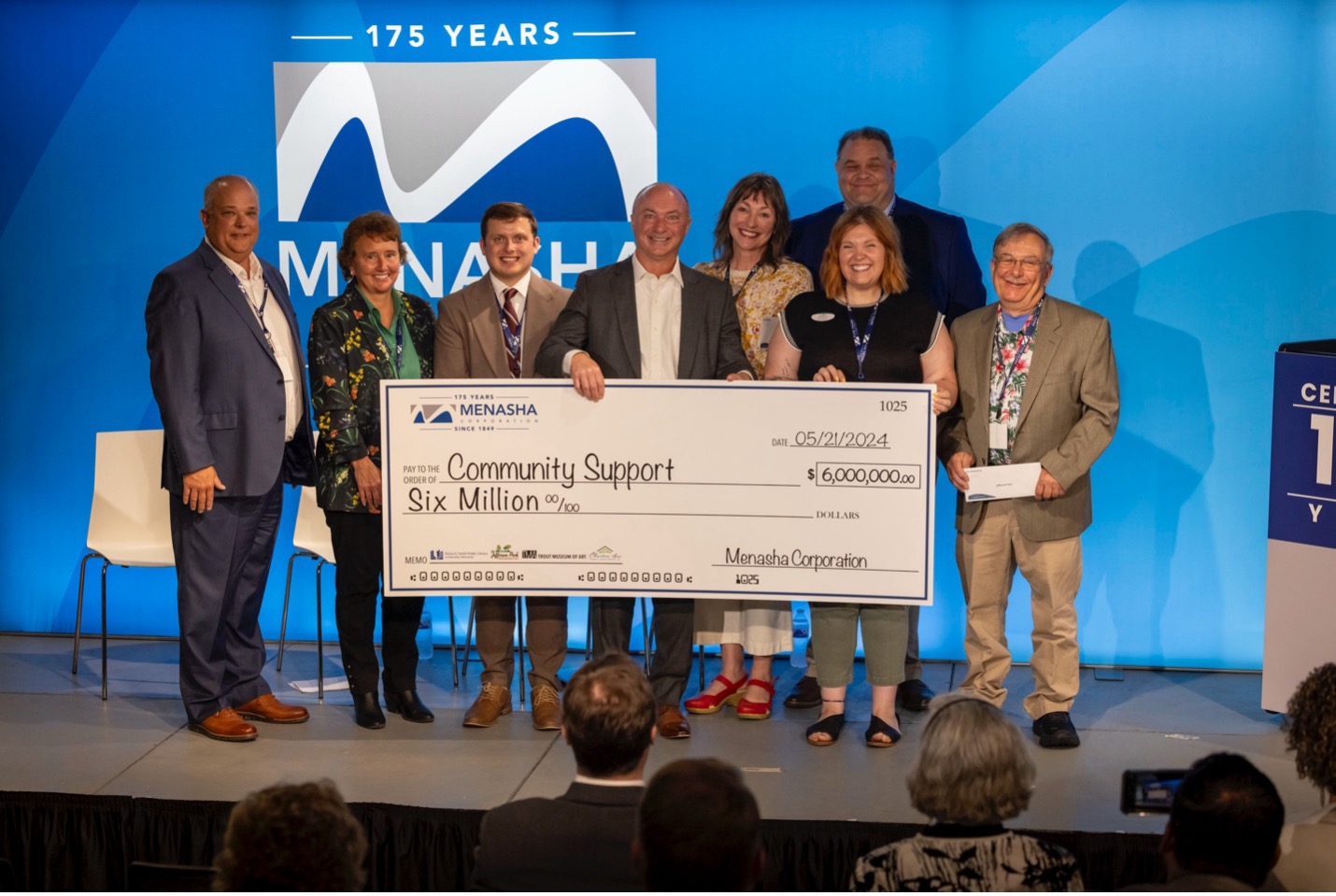 A group of nine people stands on a stage, holding an oversized check for six million dollars from Menasha Corporation for community support. A large sign behind them reads "Menasha 175 Years." The event highlights Menasha Corporation's ongoing commitment to nonprofit organizations through substantial grants.