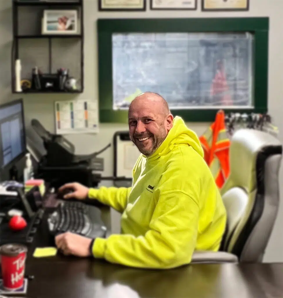A person in a bright yellow hoodie sits at a desk, smiling at the camera. The desk has a computer, keyboard, and a drink can. Wall shelves and a window are in the background.