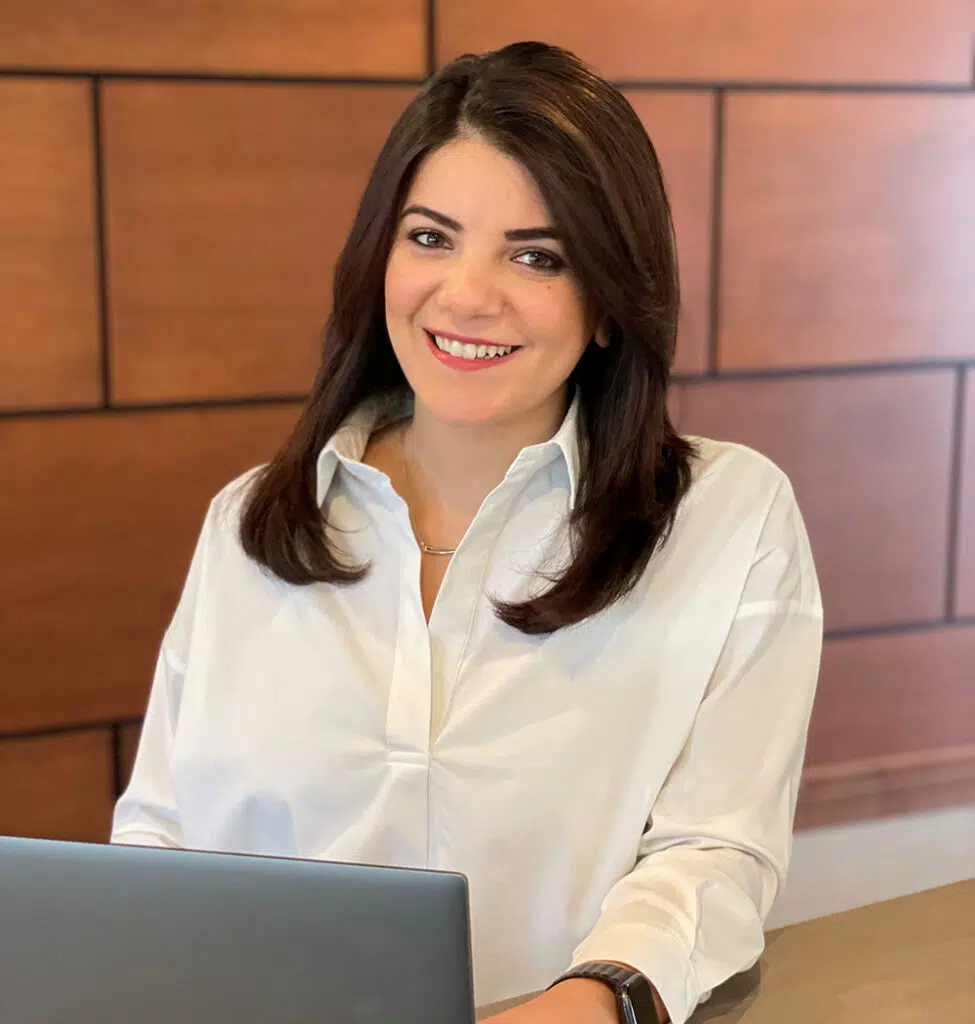Person with long dark hair, wearing a white shirt, smiling while sitting at a desk with a laptop. Wooden panel background.