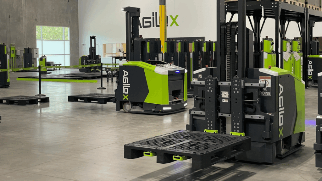 Automated guided vehicles with "Agilox" branding transport pallets across concrete floors and walls, showcasing advanced material handling in a modern industrial warehouse.