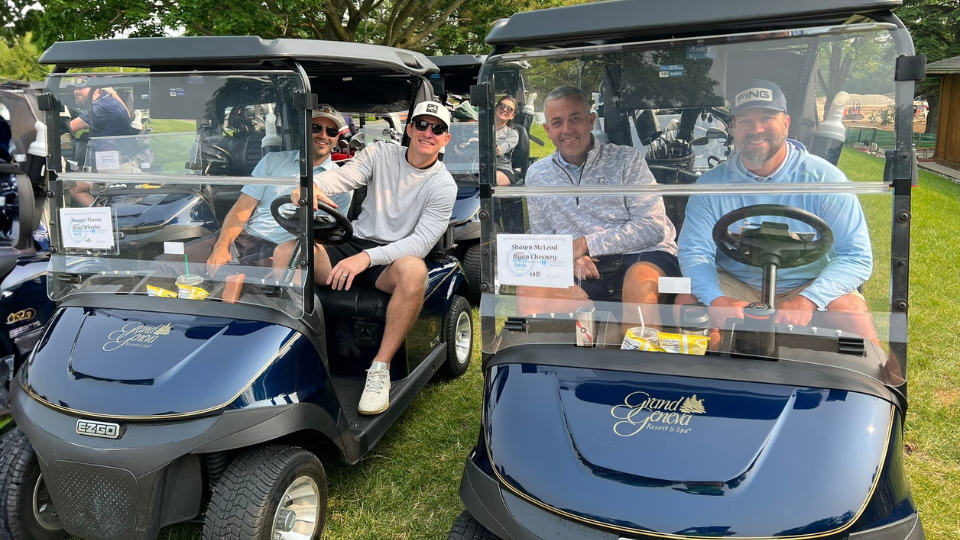 Four men sit in two black golf carts on a grassy area, smiling at the camera. Signs on the carts read "Shore School Golf Outing." It's a Good Day For Kids, with snacks visible in the carts and cheerful energy all around.