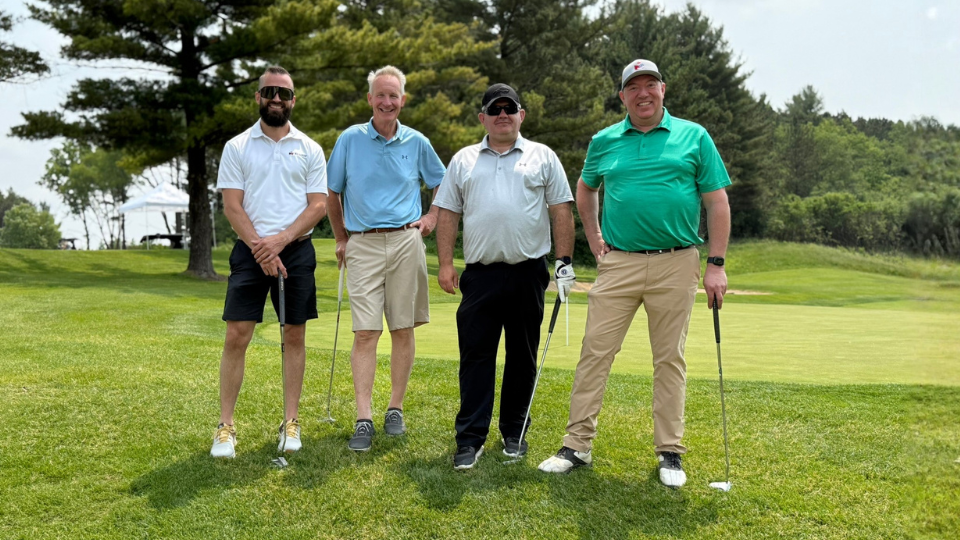 Four men stand on a golf course, each holding a golf club, enjoying Good Days For Kids among the trees and lush grass in the background.
