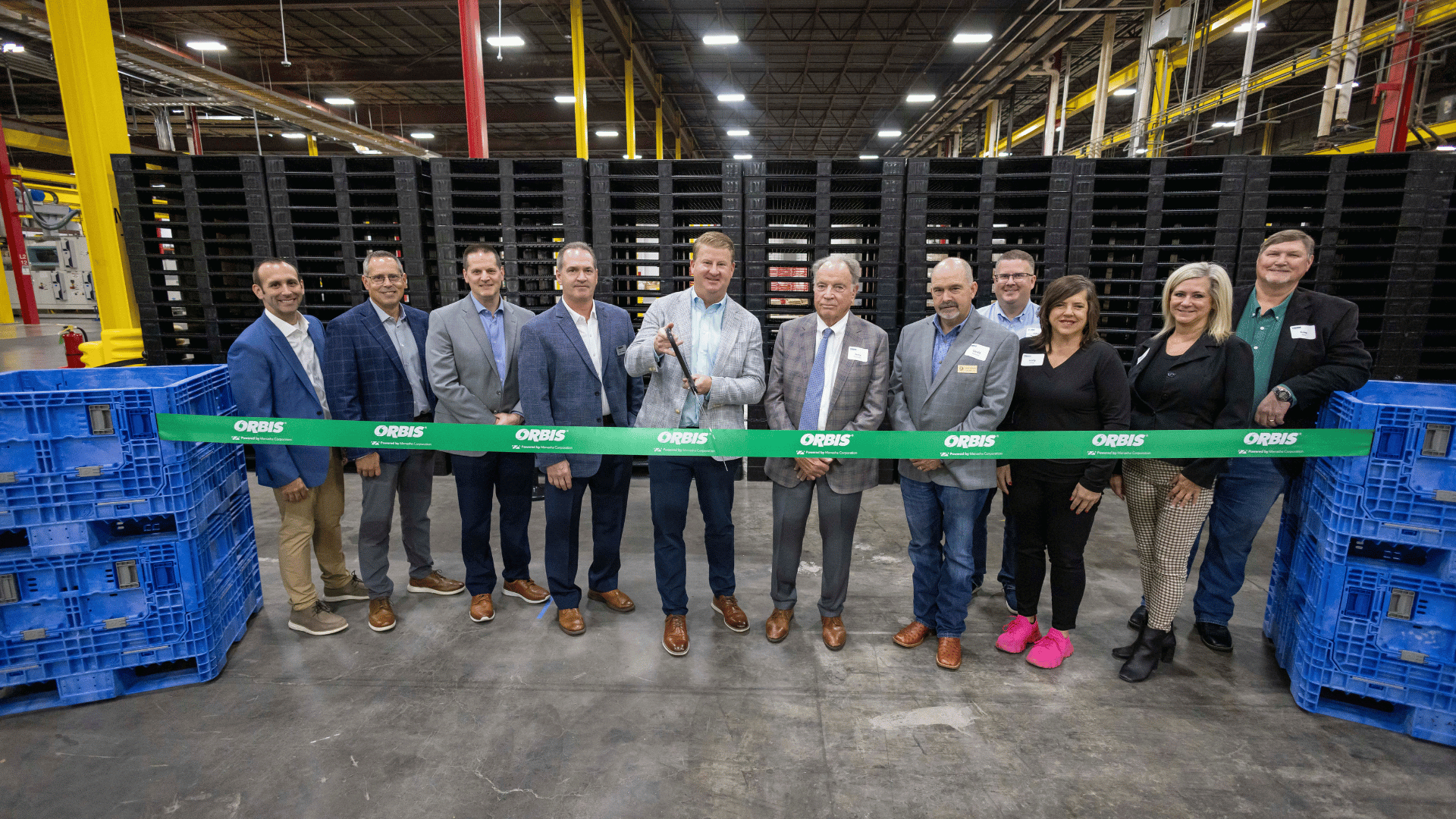 A group of people in business attire stand behind a green ribbon labeled “ORBIS” at an ORBIS Corporation manufacturing facility in Greenville, Texas, with one person holding scissors for a ribbon-cutting ceremony.