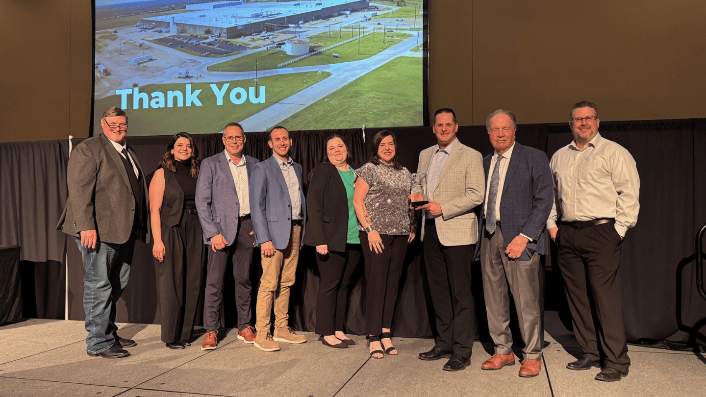 A group of ten people stands on a stage posing for a photo, some holding the Community Investment Award from the Greenville Chamber of Commerce. A "Thank You" message and aerial building image are displayed on the screen behind them.