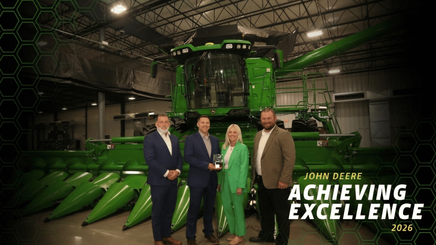 Four people in business attire stand in front of a large green John Deere combine harvester inside a warehouse. Text reads: "John Deere Achieving Excellence 2026 – ORBIS Partner Level Supplier.