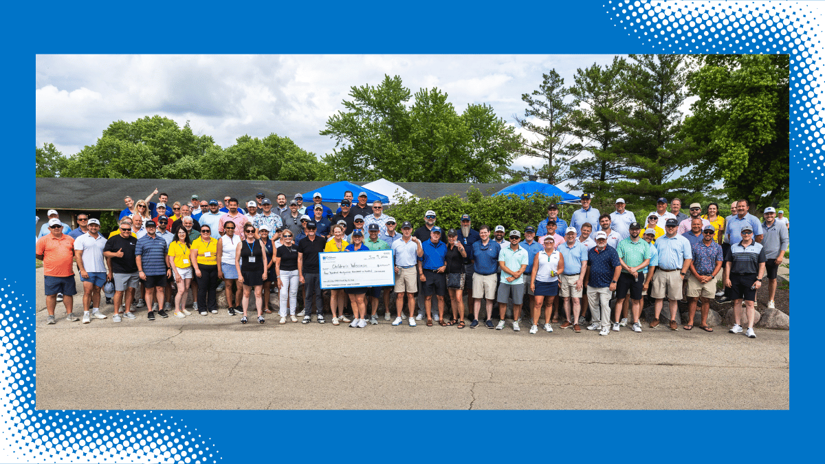 A large group of people pose outdoors, some holding a large check, with trees and a building in the background under a partly cloudy sky.