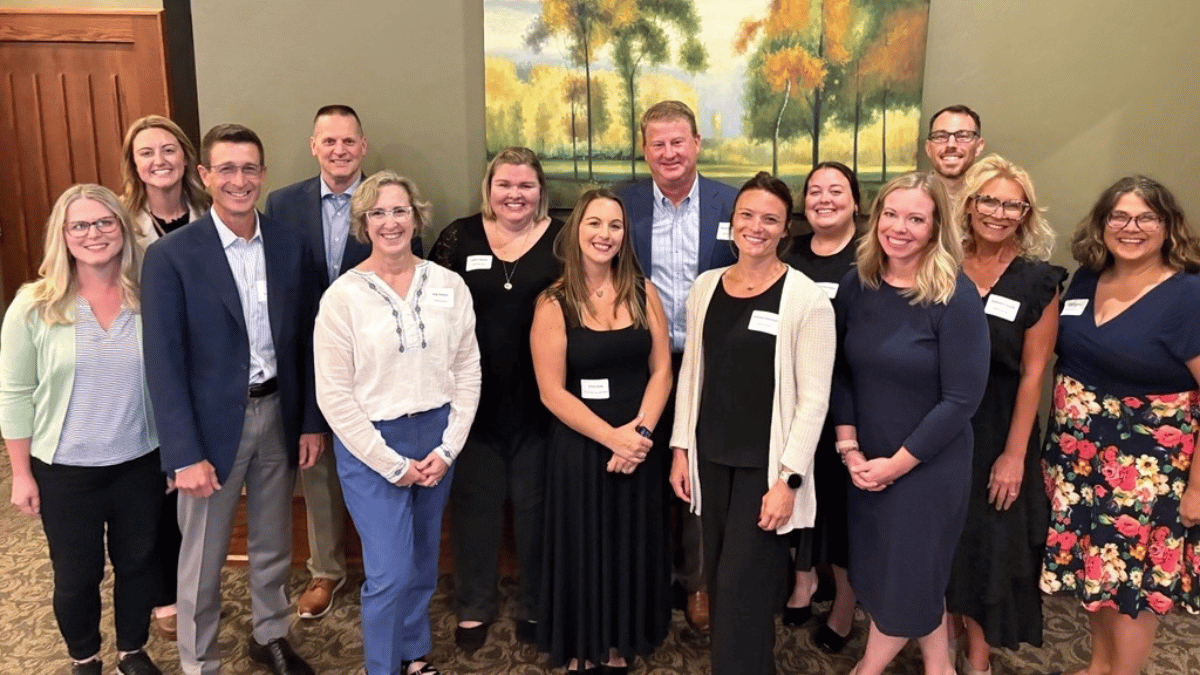 A group of thirteen adults, dressed in business casual attire, stand indoors smiling for a group photo in front of a painting of trees during the ORBIS 2026 Reception, celebrating their commitment to Community Giving.